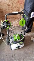 Greenworks electric pressure washer showing handle, power cord, and hose wrapped around it indoors on a concrete floor.