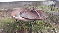 Front view of rusted steel fire pit with metal stand and damaged lid lying on top