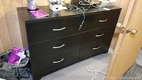 Front view of black laminate 6-drawer dresser in a basement room showing surface wear and clutter on top.