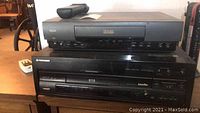 Two electronic players stacked: top is black RCA VCR tape player with remote on top, bottom is black Pioneer DVD and LaserDisc combo player.