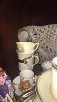 Photo showing stacked porcelain teacups with floral and bird designs inside wooden cabinet with lace doilies.