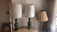 Two matching green ceramic table lamps with pleated cloth shades covered in protective plastic, placed on a dresser against the wall, alongside a mid-century modern floor lamp with linen pleated shade.