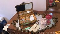 Photo showing the open ornate metal jewelry box with cherub decorations on the lid, sitting inside a metal-framed vanity tray with mirror, along with smaller trinket boxes and decorative items.