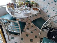 Photo showing square white-faced metal based table with several items on top (not included) and two metal chairs with green seats