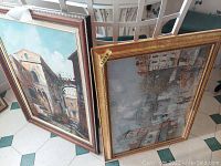Two framed paintings leaning against white chairs and a table, showcasing their fronts and frames.