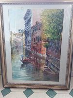 Front view of framed artwork showing gondola on canal with buildings and greenery, some glare on glass.
