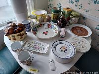 Overview of assorted ceramic serving pieces arranged on a small round table including bowls, plates, and containers with various designs and colors.