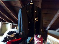 Photo showing flying cross military uniform jacket hanging with attached medals and insignia, and two feather decorated hats beneath.