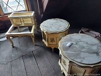 Two rectangular end tables and one hexagonal-shaped end table with cream paint, floral designs, and faux marble tops placed on wooden floor board.
