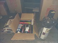 Opened cardboard box filled with assorted hardback books and magazines visible on the side, set against a wooden floor with some packing boxes in background.