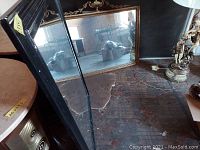 Photo showing two mirrors leaning against a wall, one with a black rectangular frame and the other with a more decorative frame, both reflecting parts of the room.