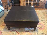 Front view of large square ottoman with dark brown leather-like top and wood base.