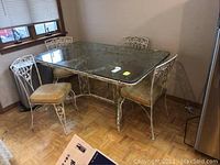 Overview of the dining table with four wrought iron chairs featuring vinyl seats. Chairs are positioned around a glass-topped iron table.