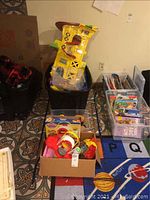 Lot overview showing bins of car racing track sets, Play-Doh kitchen playset, and boxes with assorted toys on a patterned carpet