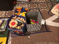 Image of fabric and plastic bins containing wooden train set parts, marble tower parts, and rubbery building toys on tiled floor.