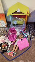 Wooden dollhouse with yellow roof, pink doll-sized beauty salon chair, Lego blocks in plastic bag, blonde doll, pink suitcase, and pouches on carpeted floor
