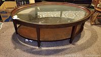 Full front-side view of oval wooden coffee table with clear removable glass top and lower shelf. Carpeted floor and patterned carpet visible through the glass.