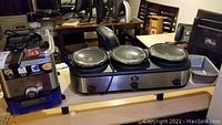 Photo shows T-Fal electric deep fryer in stainless steel with black handle and lid beside 3-well Kitchen Living crockpot unit with black lids on a wooden table.