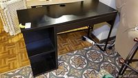 Black melamine desk from the front showing desktop, drawer, metal leg, and bookshelf base with four compartments.