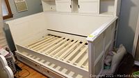 White day bed with wooden slat mattress supports and trundle storage drawer pulled out. Overhead cabinets on the back panel of the bed. Visible wear and scuffs.