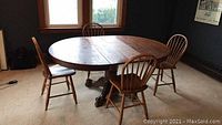 Photo of round oak dining table with leaves inserted, showing wood grain and pedestal base with four chairs around it.