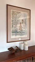 Framed winter scene picture showing snow-covered house and trees with white picket fence and lamppost, wooden frame.