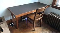 Full view of the wood desk with laminate surface and wooden chair in front of it near a window and radiator.
