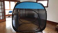 Pop up screen tent set up indoors, showing front mesh panel with zipper door, blue top and edges, and black mesh sides.