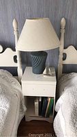 Nightstand with drawer, open shelf holding several hardcover books, blue ceramic lamp with cream fabric shade, and silver sailboat figurine on top, positioned between two headboards.