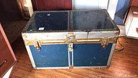Front and top angled view of antique trunk showing blue painted panels with silver metal framing and corner protectors. The trunk appears aged with scratches and wear marks.