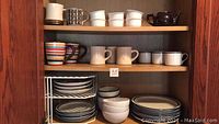 Shelves containing various stacked plates, bowls, and mugs of different styles and colors, including striped and patterned mugs.