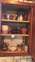 Photo of open wooden cabinet shelf showing a metallic teapot with black handle on top shelf with a small metal bowl. Middle shelf shows a beige-beige-brown mug, three dark brown glass cups, and a pink ceramic bowl. Bottom shelf contains a small brown ceramic pitcher, cream-colored bowl with scalloped edges, two small ceramic mugs, and a small textured cup.