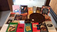 Image showing assortment of 13 cookbooks, a round wood platter and a wooden box on a table.