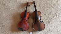 Photo showing two antique violins side by side from front view, resting on carpet, missing strings