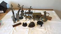 Full view of aluminum figurines, horse and wagon, and brass items on white cloth including two aluminum deer silhouettes, musician figures, horse figurine, brass pitcher, and small brass decorative objects.