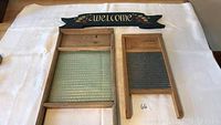 Two antique washboards and two painted wooden welcome signs displayed on white cloth.
