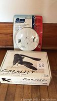 Photo of Corklift wine opener in box and Garrison smoke and carbon monoxide alarm in packaging on wooden floor.