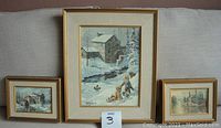 Set of three framed Keirstead art prints displayed together on a sofa showing their relative sizes and themes of snowy winter scenes with houses and figures.