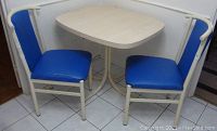Photo showing full kitchen table and two chairs in corner, cream-colored table top and metal frames with blue vinyl upholstery on chairs