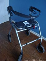 Silver adjustable walker with black seat and hand brakes against a blue wall on wood floor.