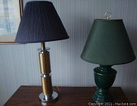 Two table lamps on a wooden surface with one lamp having a gold-tone base and dark blue shade, the other with a green ceramic base and dark green shade.