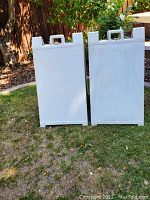 Two white plastic A-frame signs displayed standing side by side on grass outside.