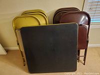 View of folded card table and 8 folding chairs stacked against wall, showing table top and chair backs.