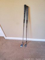 Two golf wedge clubs standing against a wall showing their full length and grips.