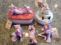 Five Pink Panther themed ceramic pieces including a music box, storage box with lounging Pink Panther lid, and three Pink Panther character figurines in different poses on a stone countertop.