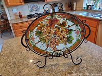 Front view of oval stained glass server on metal stand, showing grapevine motif with clusters of grapes and leaves in shades of green and purple with brown borders.