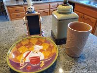 Photo showing four kitchen items on countertop: chef chalkboard figure, serving bowl with chef print, green ceramic canister, peach ceramic wine cooler