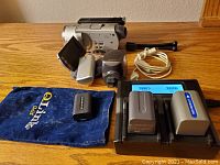 Sony DCR-SR100 Handycam video camera, multiple batteries, charger, cables, pocket stand, soft carrying case for microphone, Bluetooth mic units on wooden surface