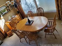 Overall view of the oak oval dining table with six chairs arranged around it, in a carpeted room near windows with curtains.