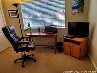 Full view of office desk setup by window with black leather adjustable chair and floor desk lamp, side TV table with TV on top